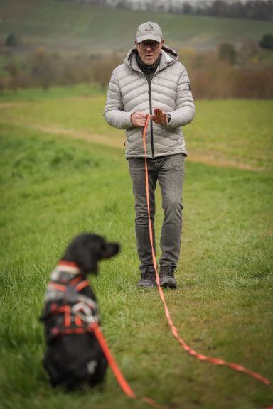 Mann mit grauem Hut und Jacke hält eine Leine, schwarzer Hund sitzt neben ihm auf dem Gras.