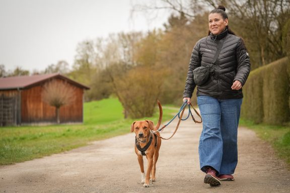 Frau mit einem Hund an der Leine, spaziert auf einem Weg in der Natur.
