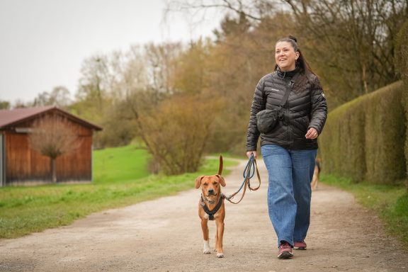 Frau geht mit einem Hund auf einem Weg entlang von Bäumen und einem Holzhaus.