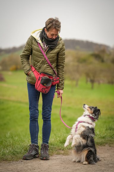 Frau mit Hund an der Leine, beide stehen auf einem Weg umgeben von Wiese und Bäumen.