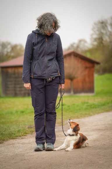 Frau mit grauen Haaren steht an einem Weg, ein kleiner Hund sitzt davor.