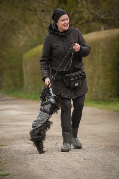 Eine Person in schwarz mit einem Hund an der Leine geht einen Weg entlang.