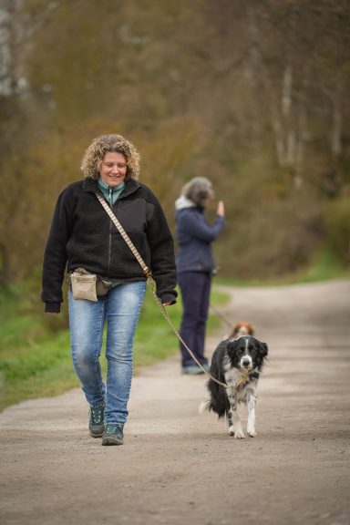 Frau mit Locken geht mit einem Hund einen Weg entlang, eine andere Person im Hintergrund.