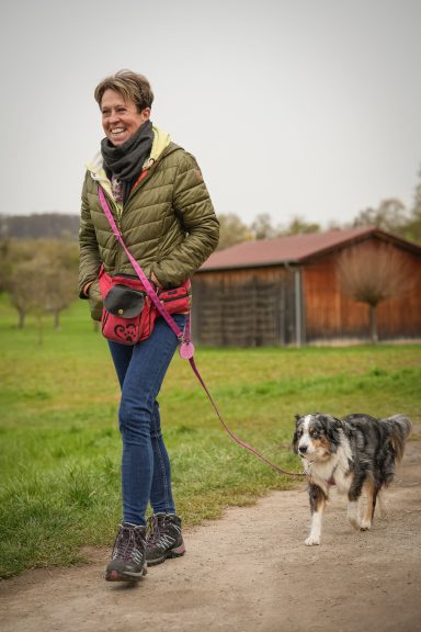 Frau mit Hund geht einen Weg entlang, umgeben von Wiesen und einem Holzschuppen im Hintergrund.